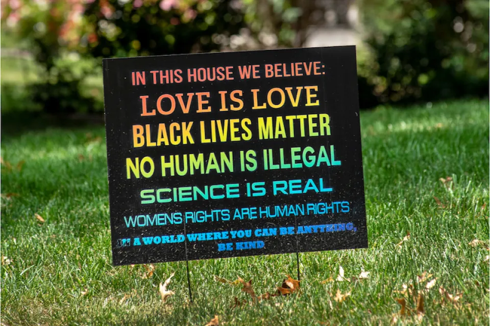 A rainbow colored sign planted in the front yard of a typical suburban home reads, "In this house we believe love is love, black lives matter, no human is illegal, science is real, women's rights are human rights. In a world where you can be anything, be kind."